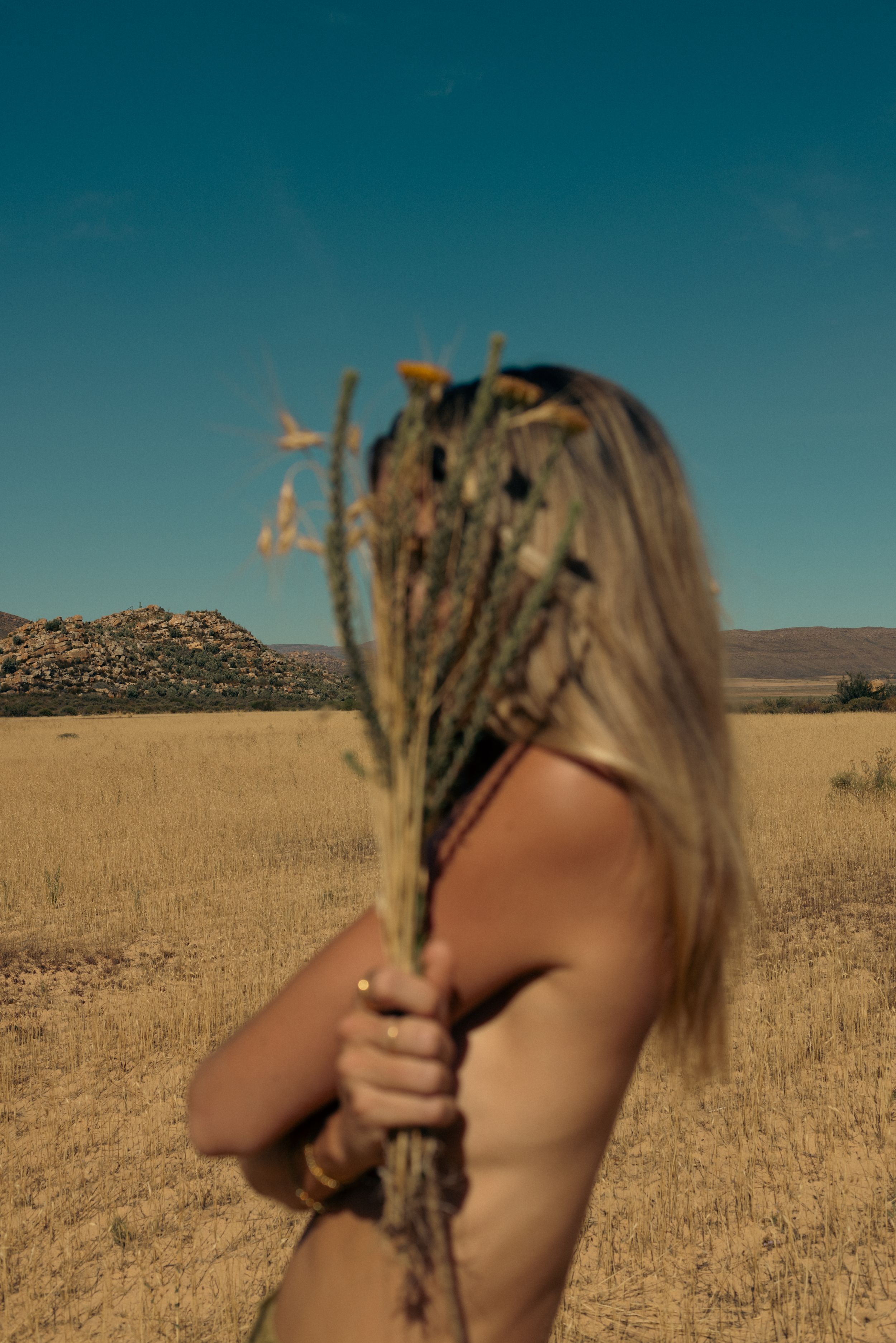 A person holding dried plants covers their face in a sun-dried grassland landscape.