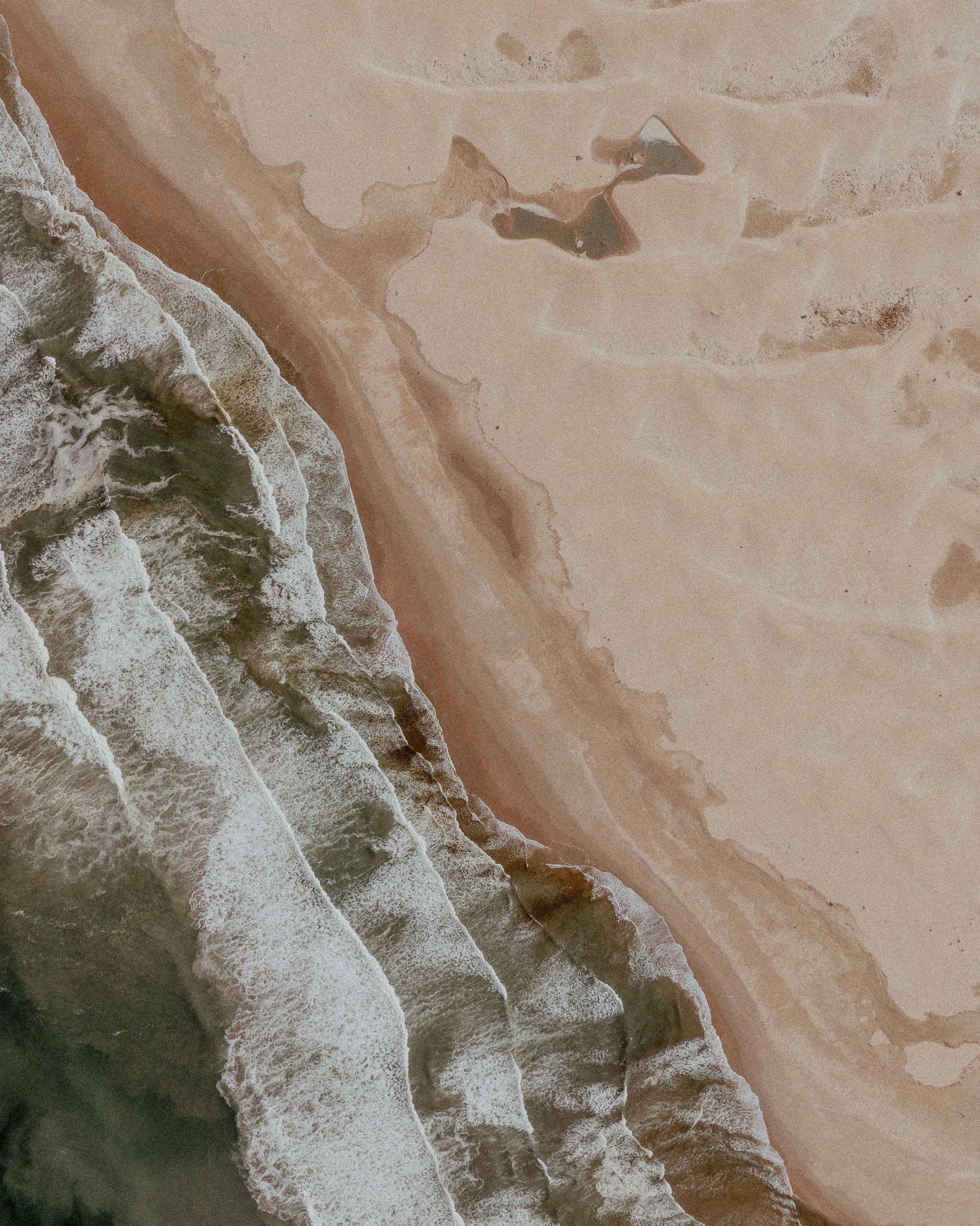 Aerial view of coastline showing waves meeting sandy beach in muted earth tones.