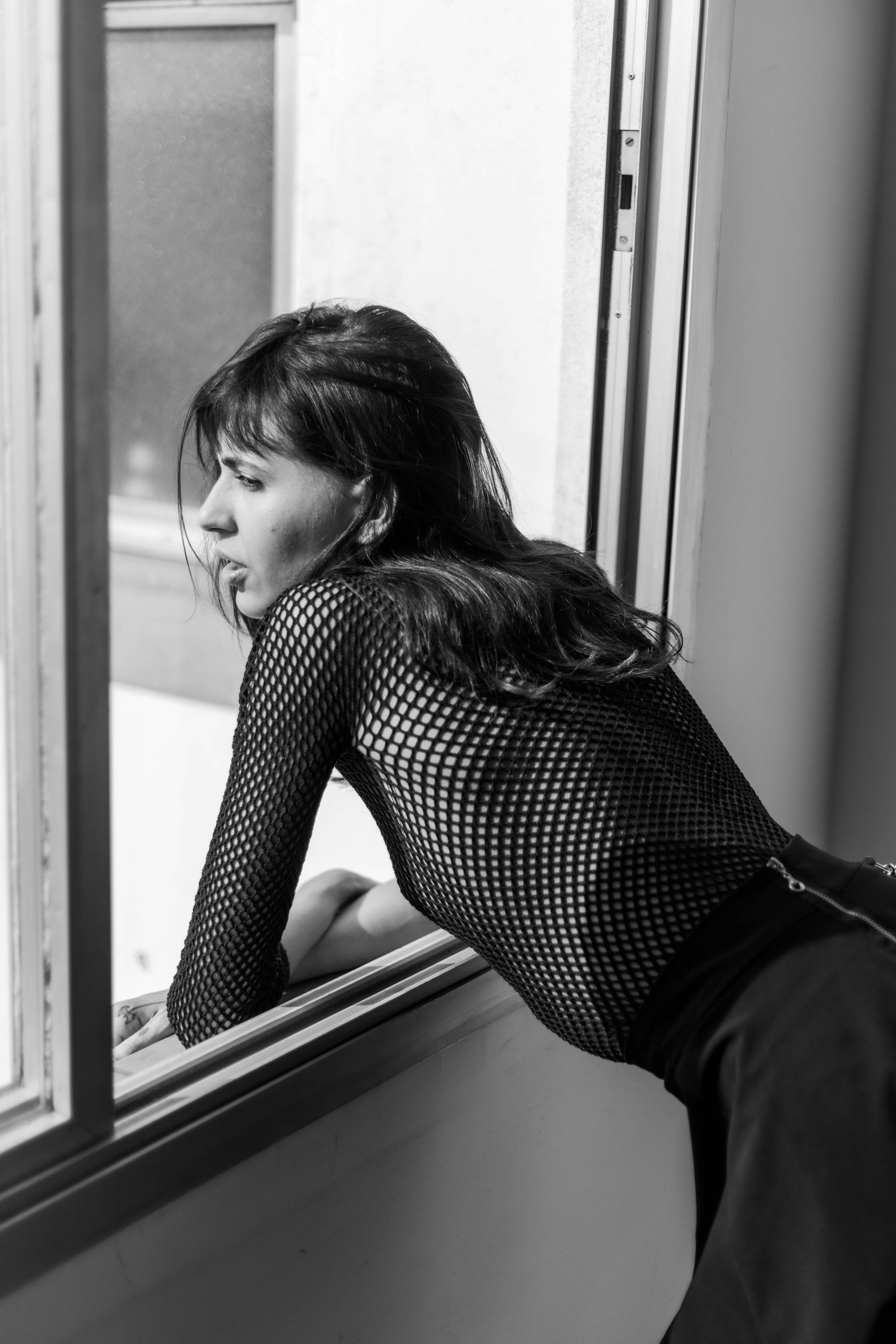 Black and white portrait of someone in a mesh top looking out a window in dramatic lighting.