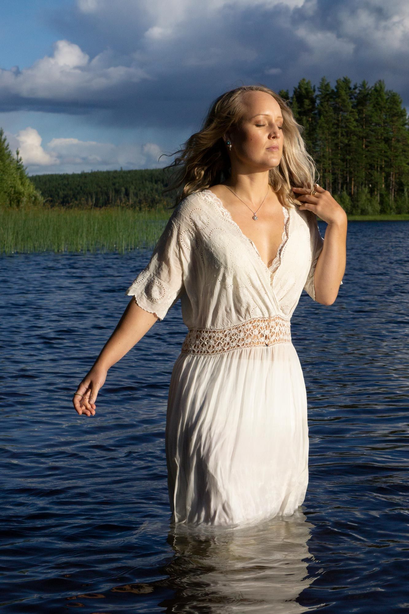 En blond kvinna som står i vattnet i en sjö en solig sommardag. Hon har på sig en vit klänning.