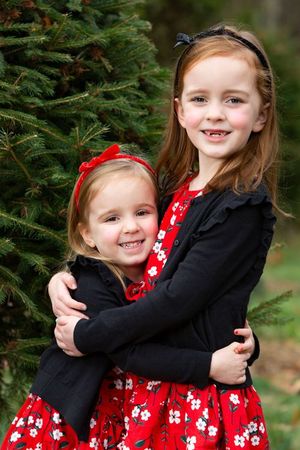 Two young girls in matching red floral dresses and black cardigans hugging outdoors near a green pine tree.