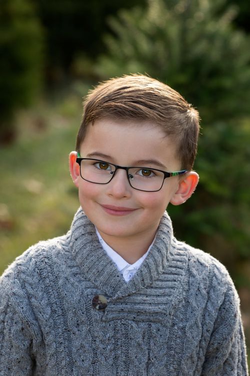 A young boy wearing glasses and a gray knit sweater smiles outdoors near evergreen trees.