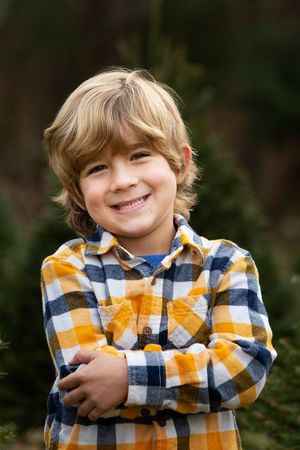 Smiling blond boy in plaid shirt with arms crossed, posing outdoors near evergreen trees.