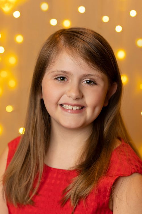 Smiling young girl with long brown hair wearing a red dress posed against a warm bokeh light background.
