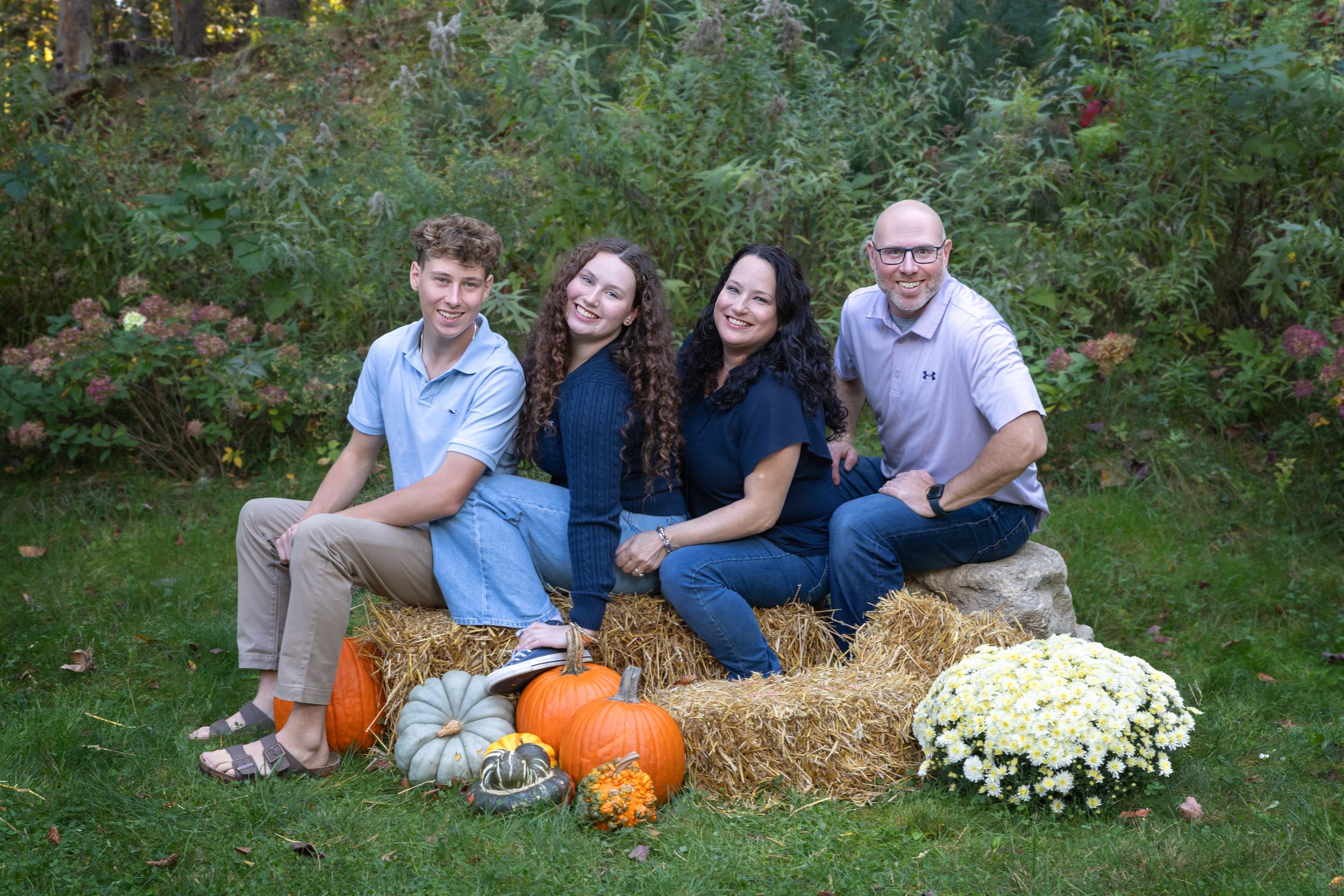 Family of four smiling outdoors in fall setting with hay bales, pumpkins, and mums for autumn photo session.