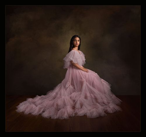 Woman in elegant pink tulle ball gown posed gracefully on wooden floor against moody studio backdrop.