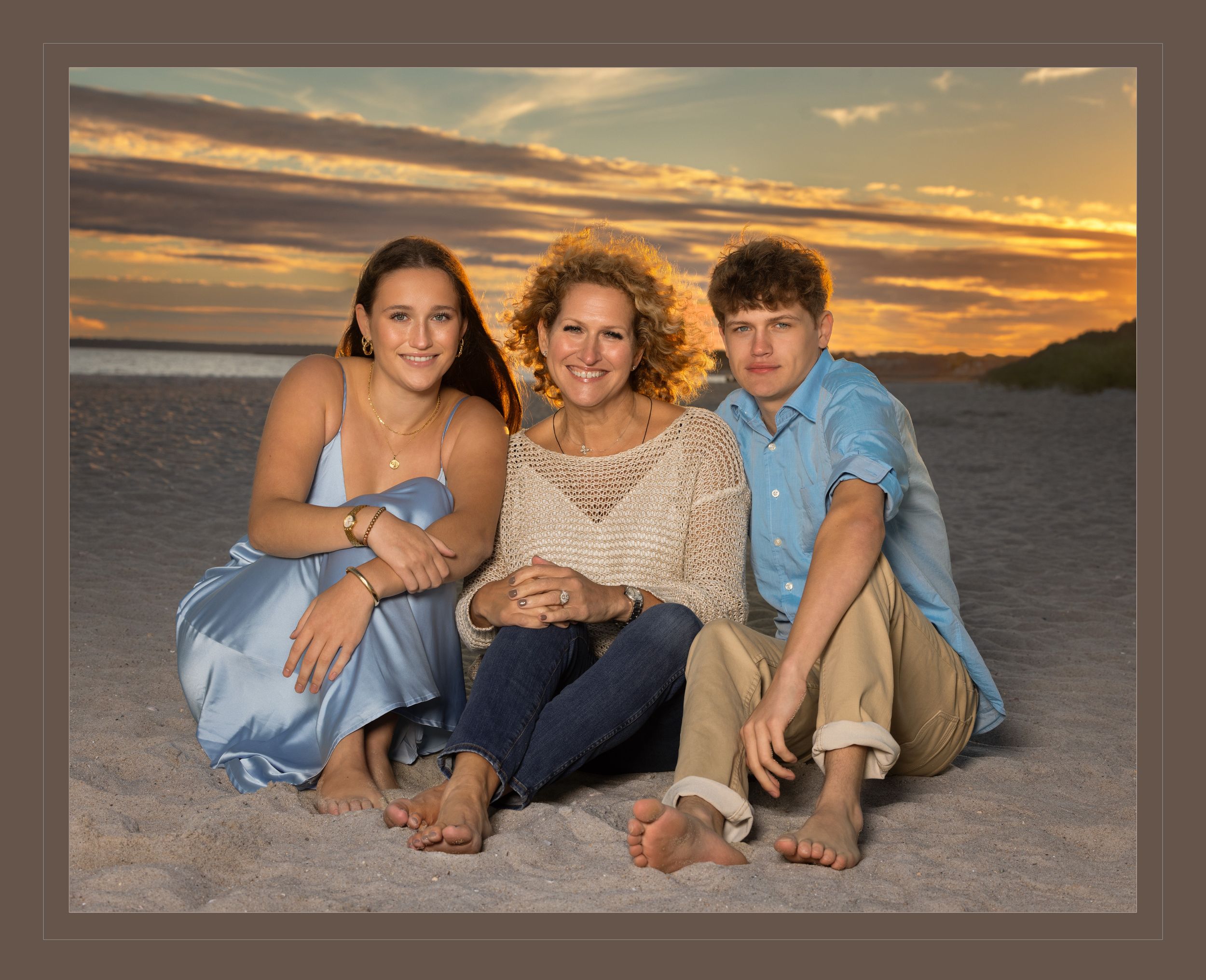 Family of three posing together on sandy beach during stunning golden sunset, smiling warmly.