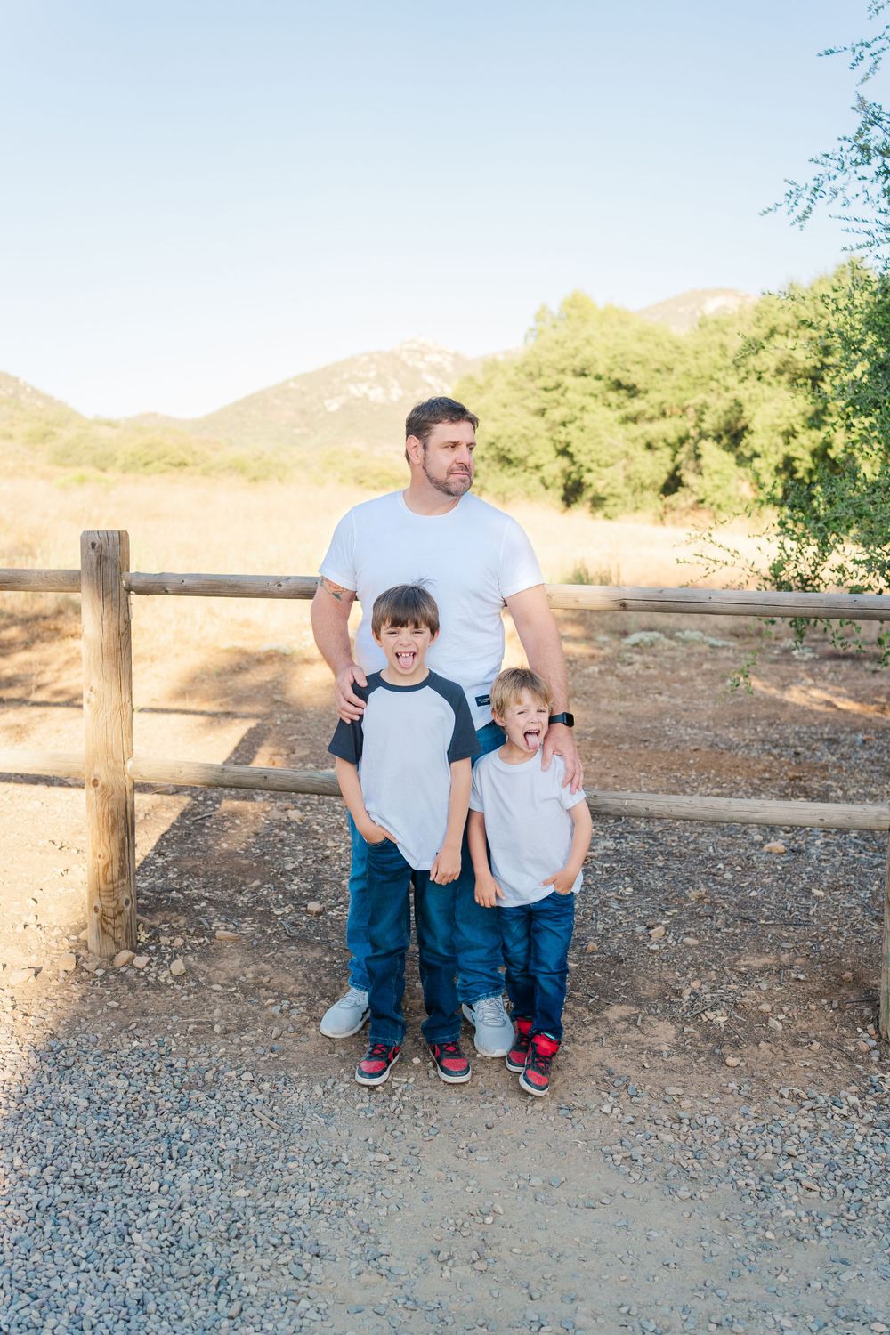 Three people standing together on a dirt path by a wooden fence with mountains in the background during sunset.