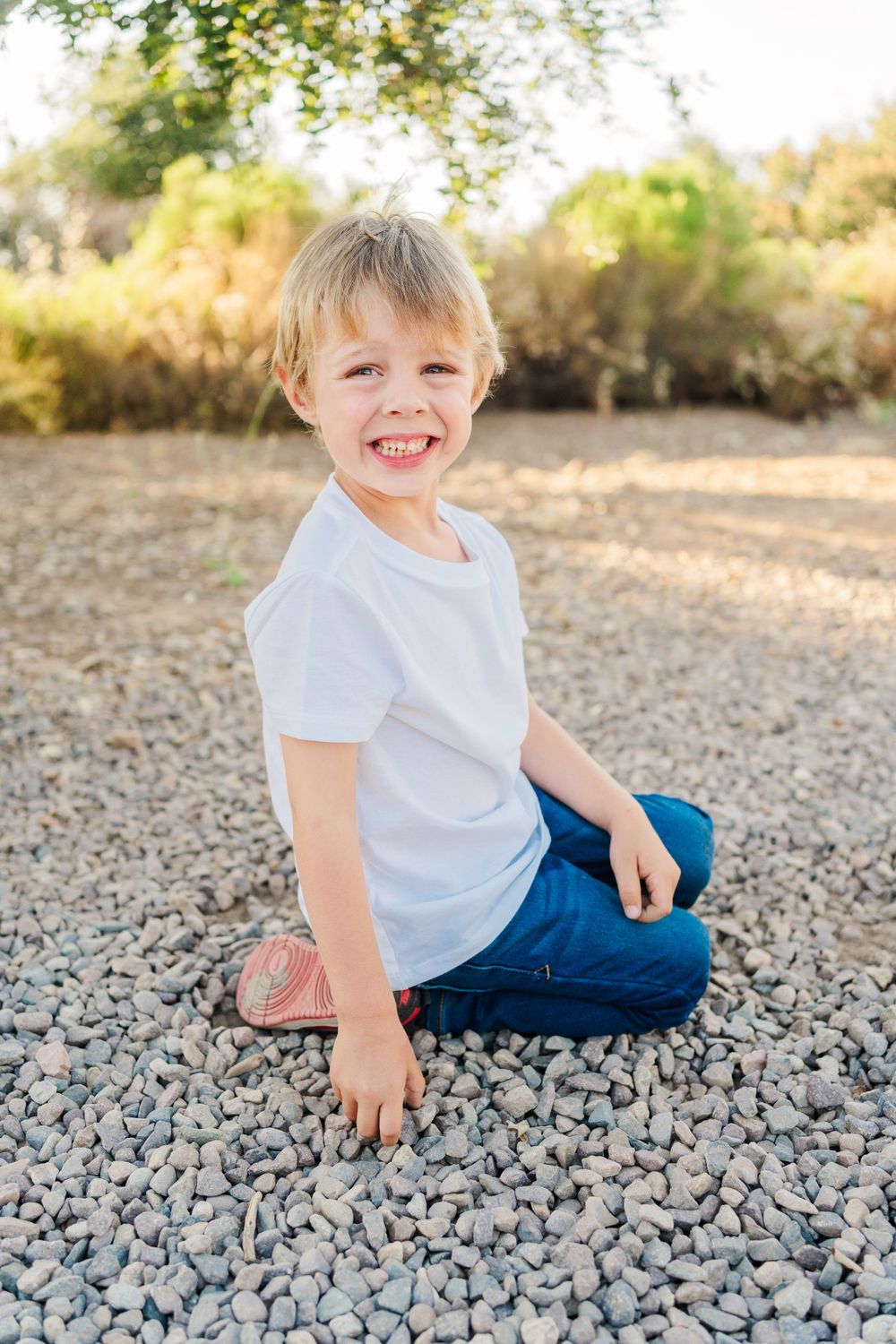 Child in white t-shirt and blue jeans sits smiling on rocky ground in sunny outdoor setting.