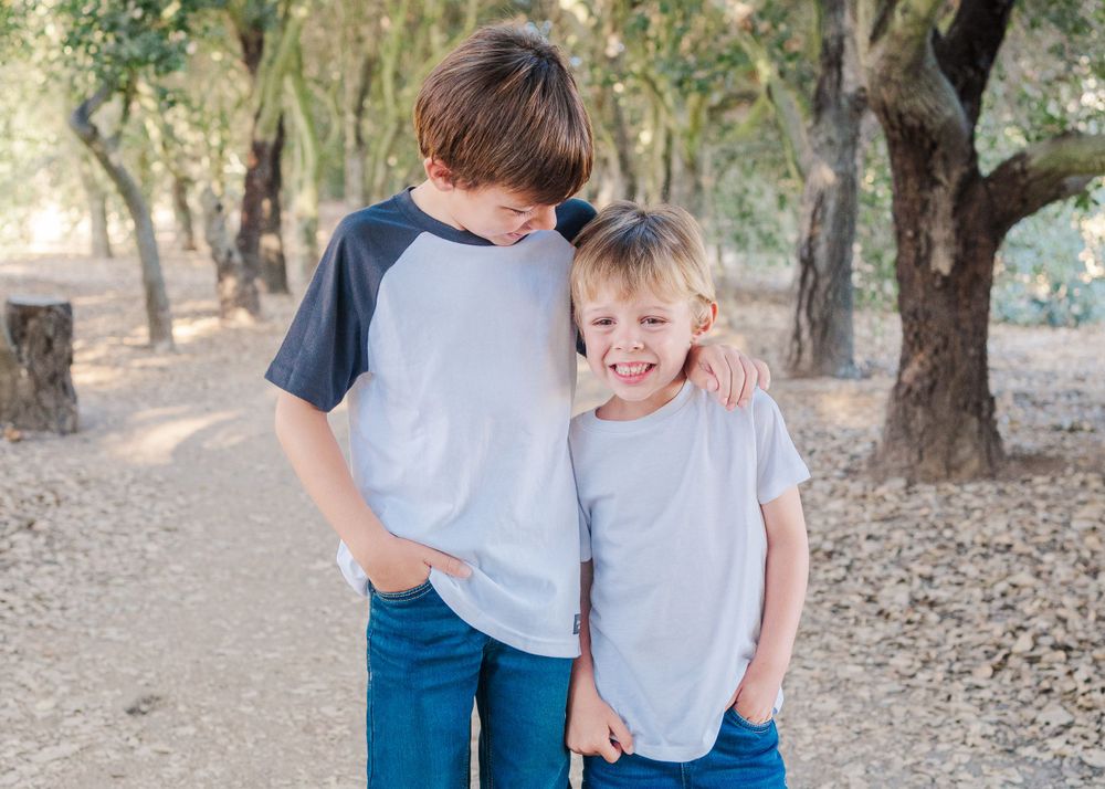 Two children wearing casual t-shirts share a sweet moment together while standing in a wooded outdoor setting.