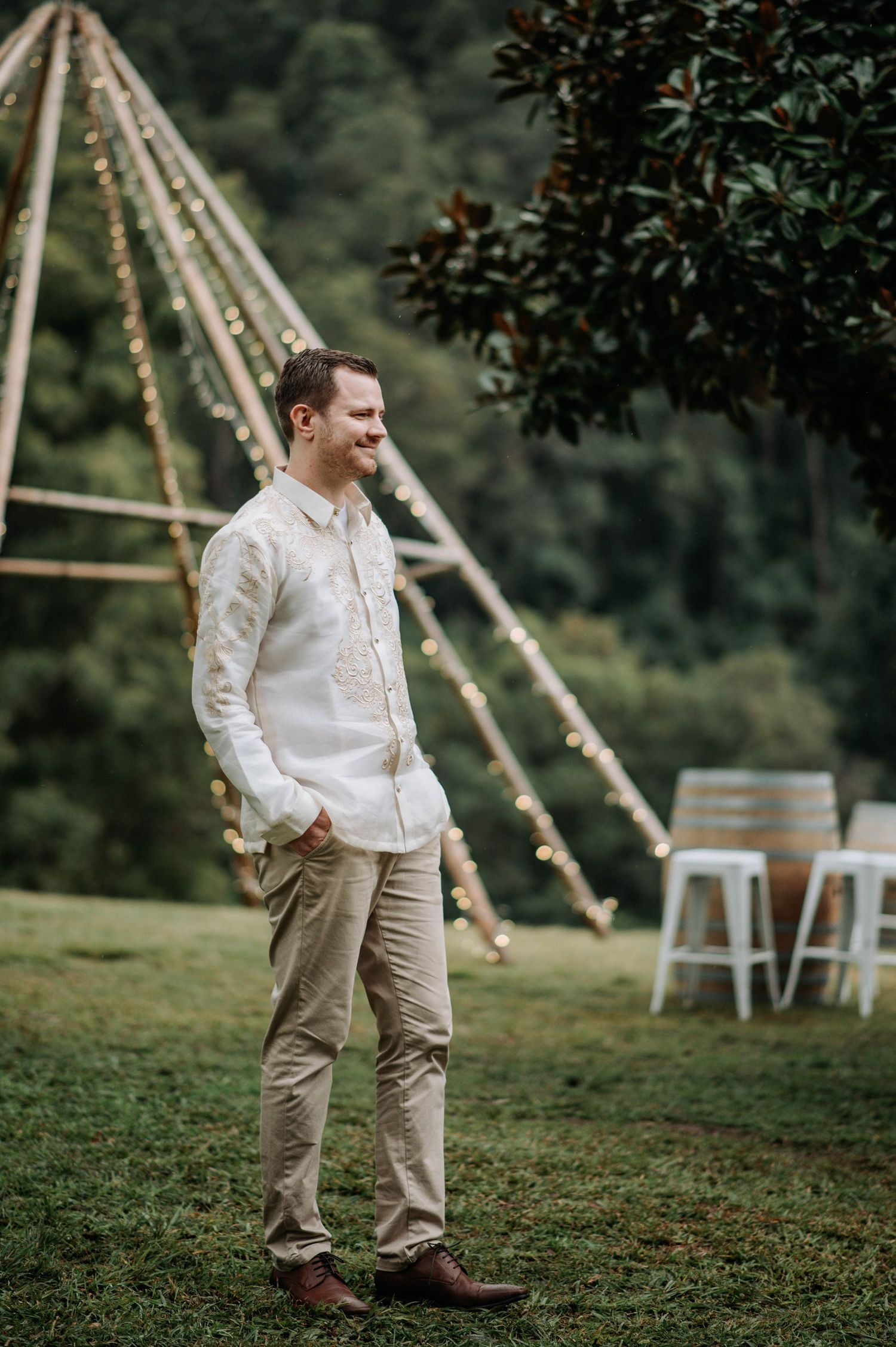A groom in traditional Filipino attire stands near a decorated teepee structure at an outdoor venue.