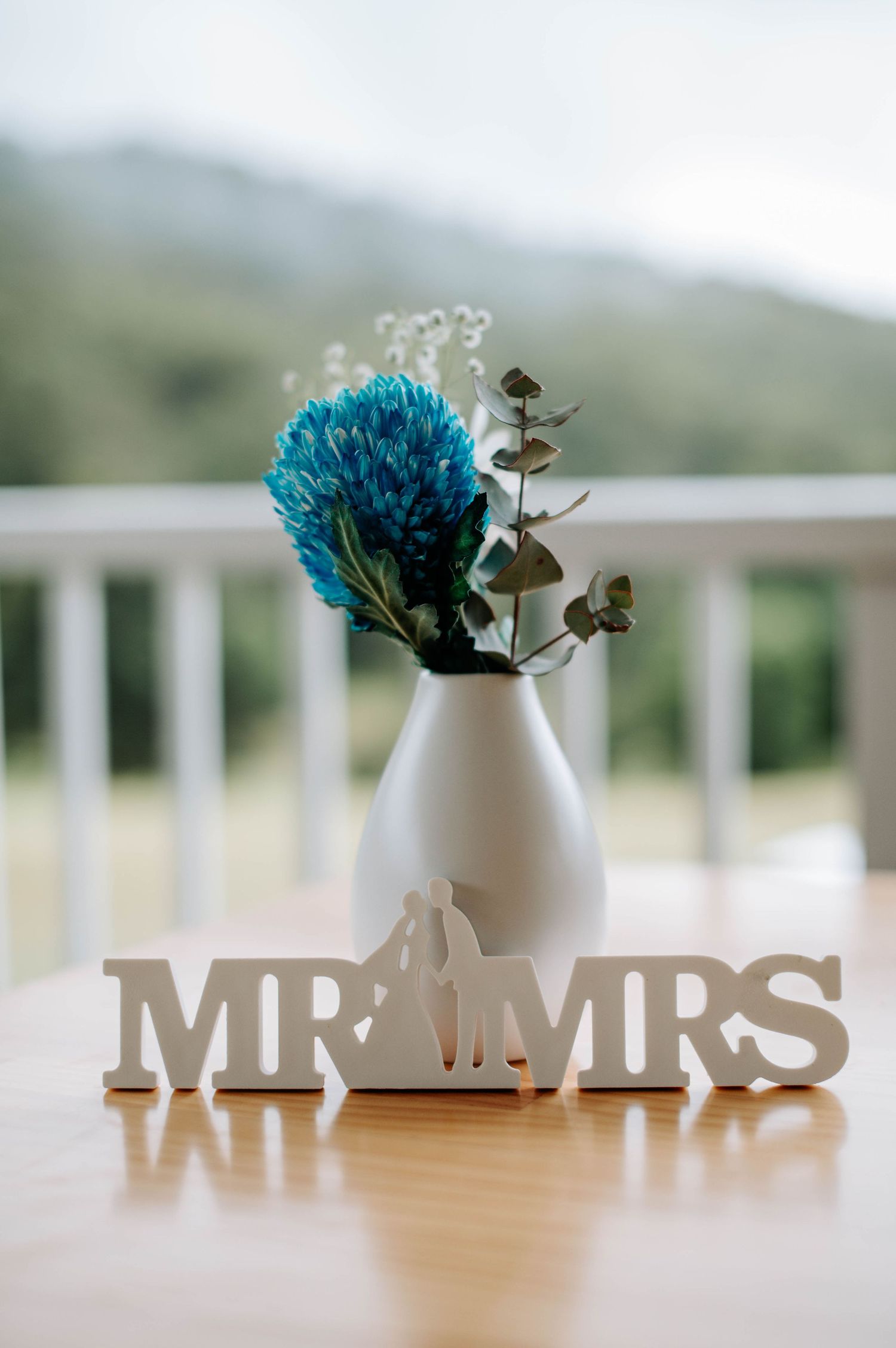 White ceramic vase with blue flower and 'MR & MRS' wooden letters on table.