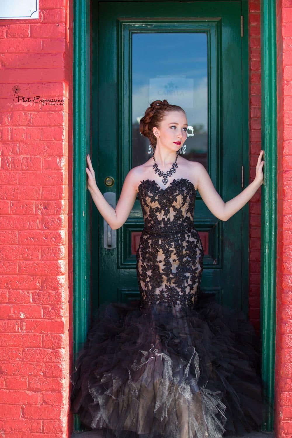 A dramatic black lace dress is showcased against a vibrant red brick wall with green doorframes.