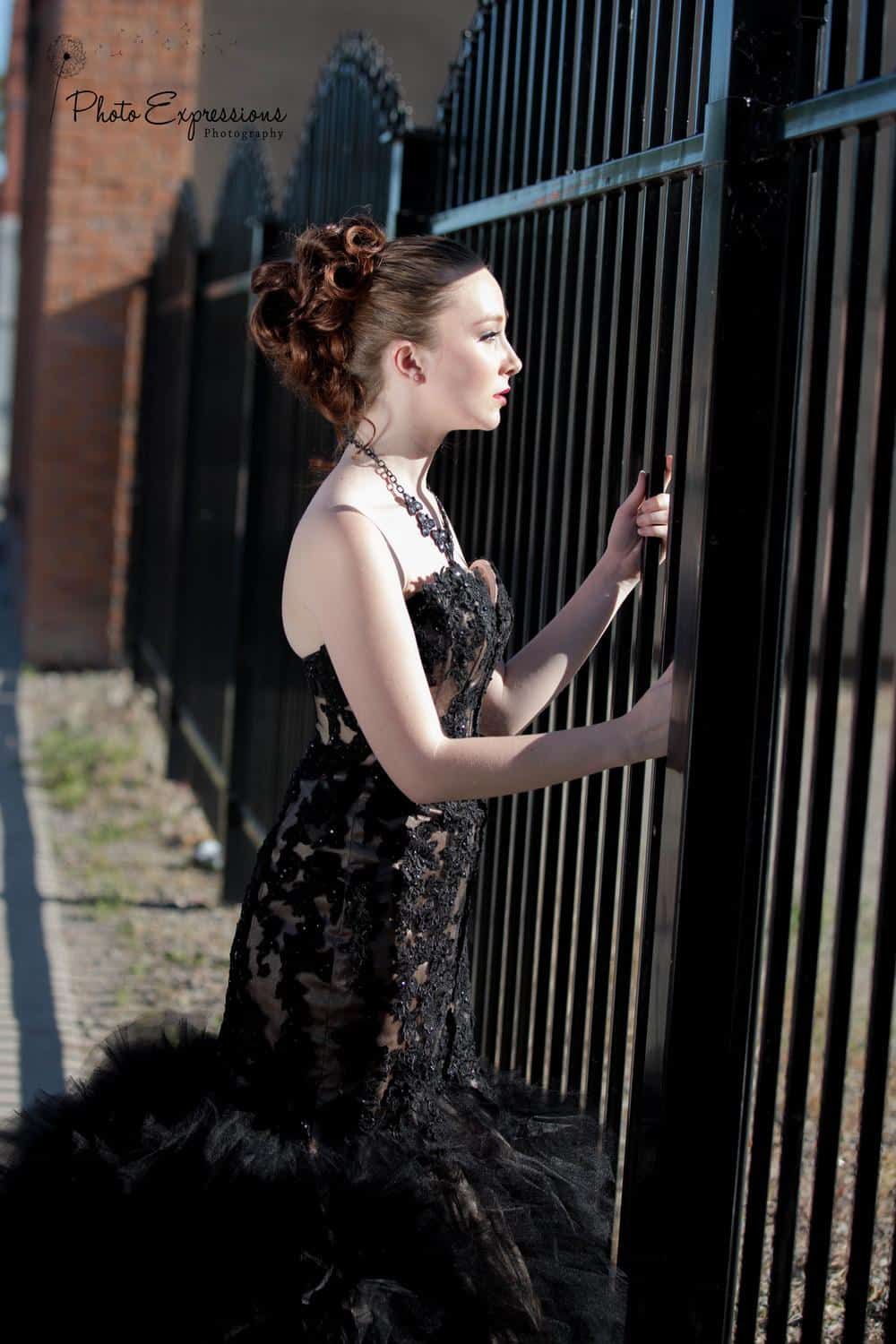 A model in a black lace gown poses dramatically against industrial metal railings in an urban setting.
