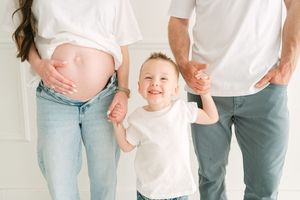 Child in white shirt holds hands with parents while standing between them, for maternity photography in an Edmond, OK Studio