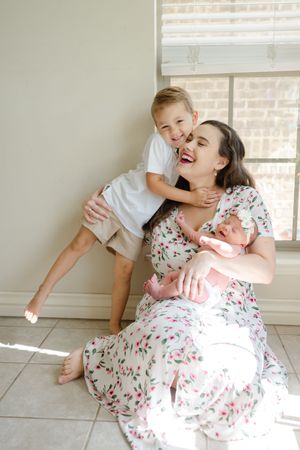 mother sits in front of window embracing older child while holding newborn for Lindsey K Brown Photography, newborn photographer