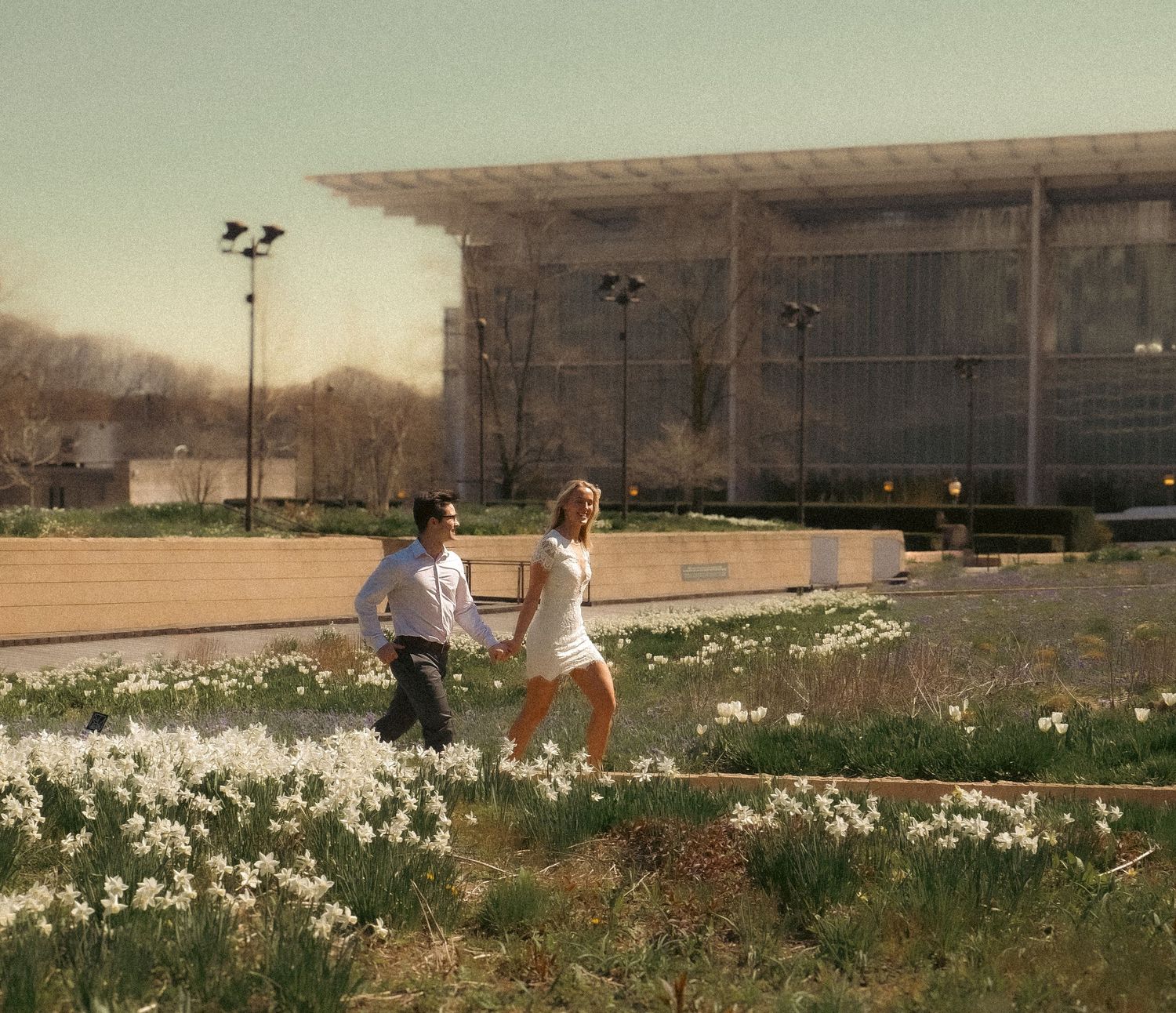 Couple runs through a field of daffodils near a modern building in vintage-toned lighting.