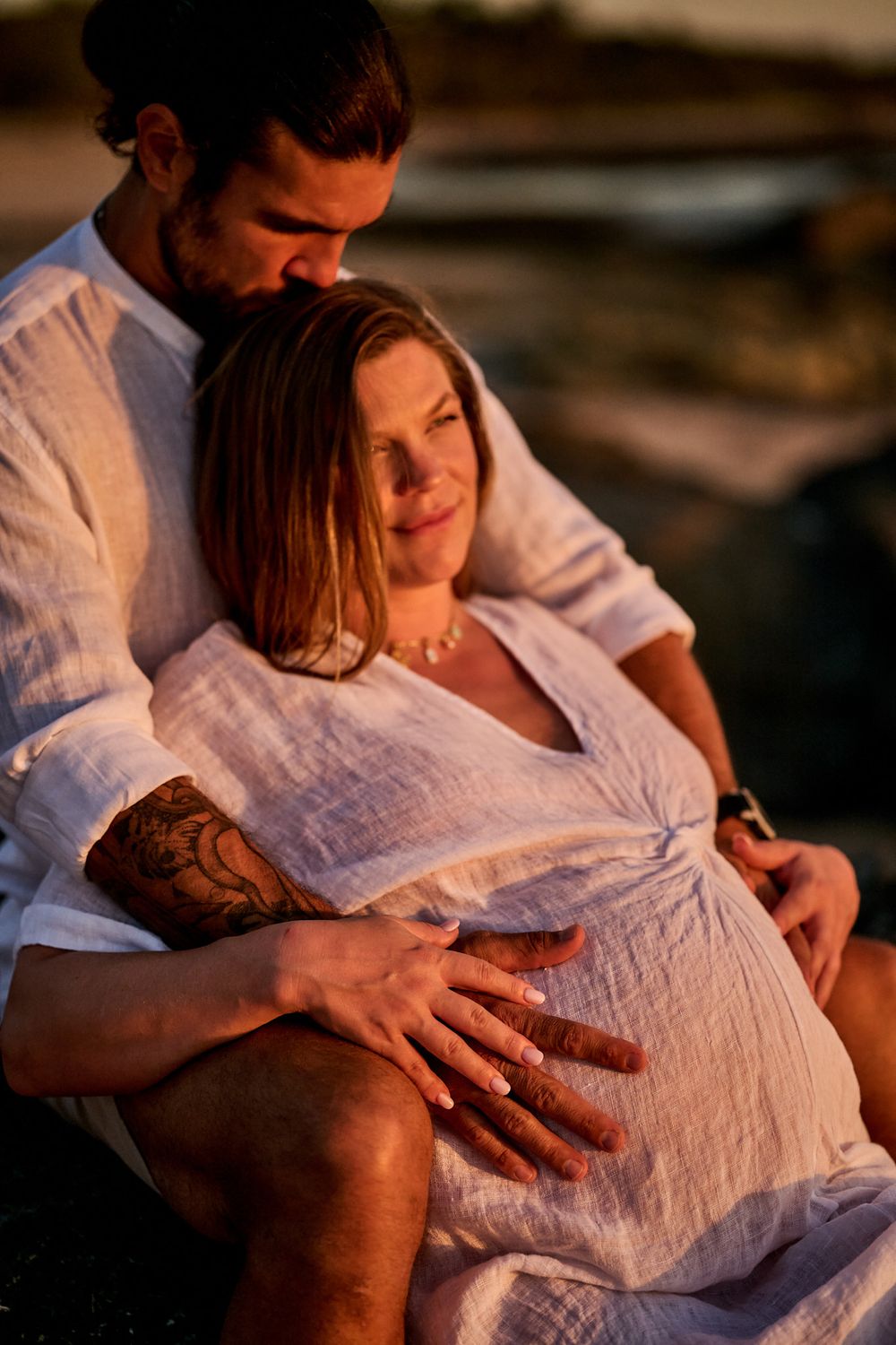 Couple in white linen sharing a tender maternity moment at golden hour, man embracing pregnant woman from behind on beach.