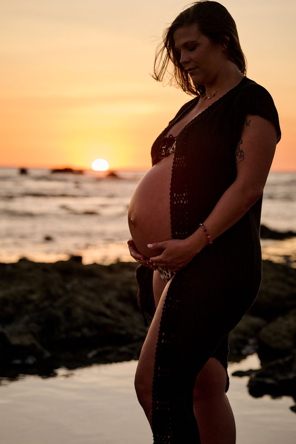 Pregnant woman in black dress cradling her baby bump on rocky beach at golden sunset.