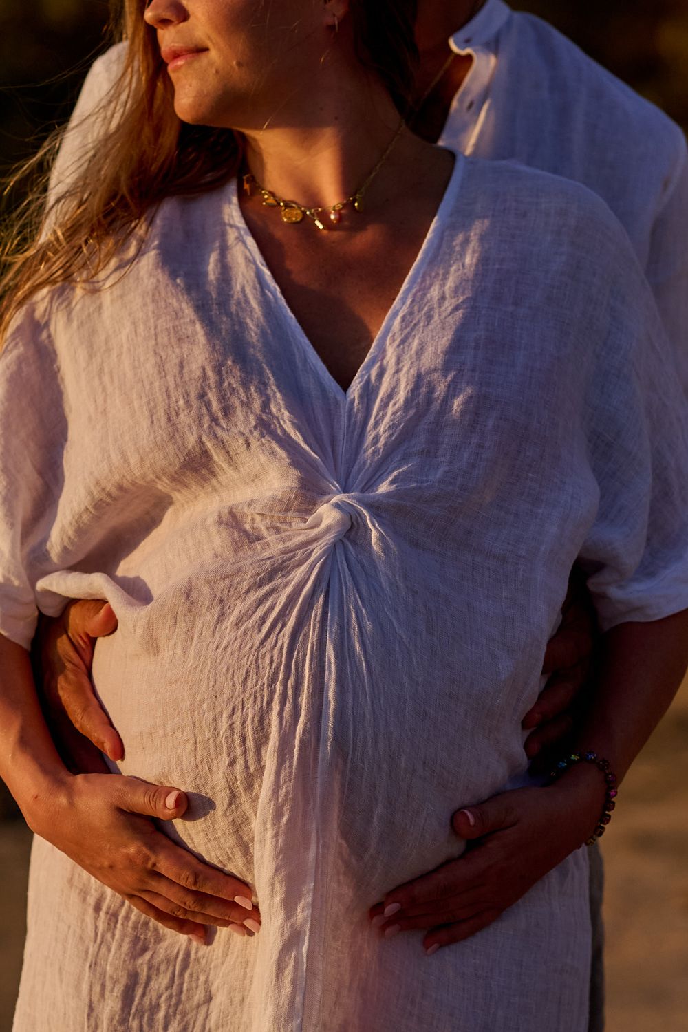 Pregnant woman in white linen dress cradled by partner's hands at golden hour sunset.