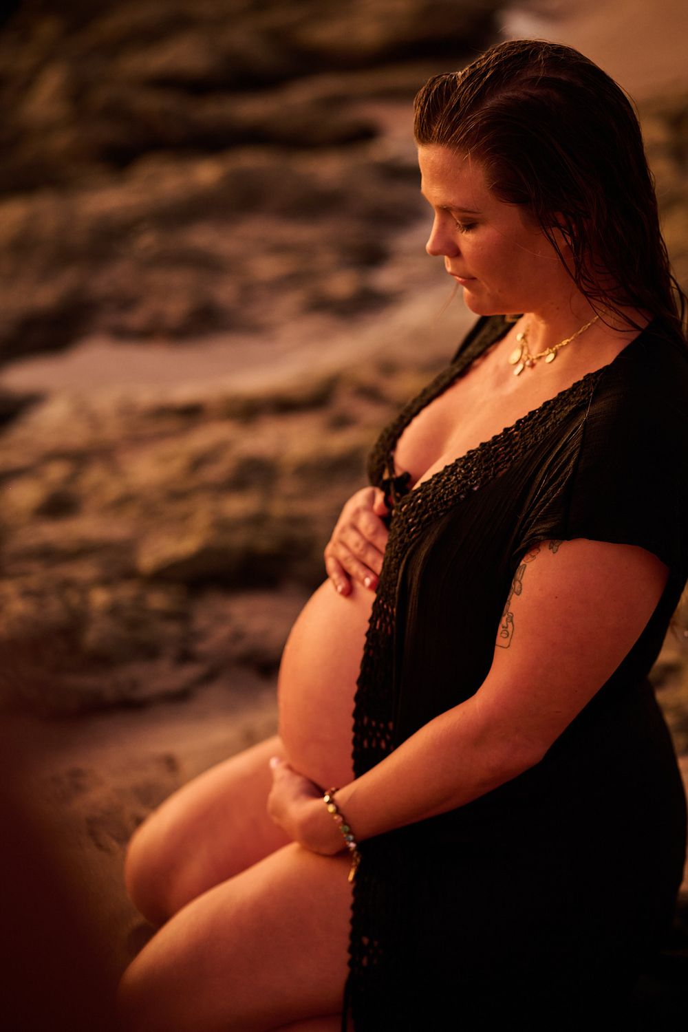 Pregnant woman in black lace dress kneeling on beach rocks at sunset, cradling her baby bump with both hands.