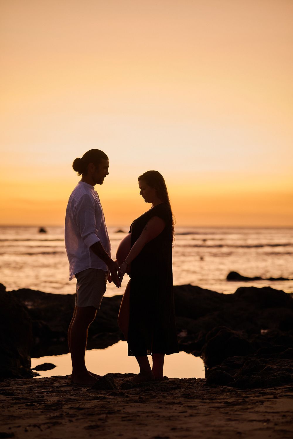 Silhouette of a couple holding hands on a rocky beach at sunset with golden orange sky.