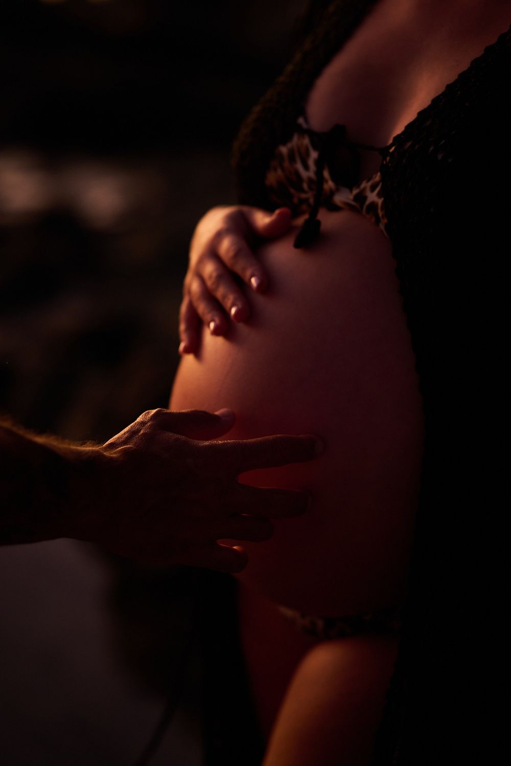 Close-up of a pregnant woman's bare belly being gently cradled by two hands in warm, dramatic lighting.
