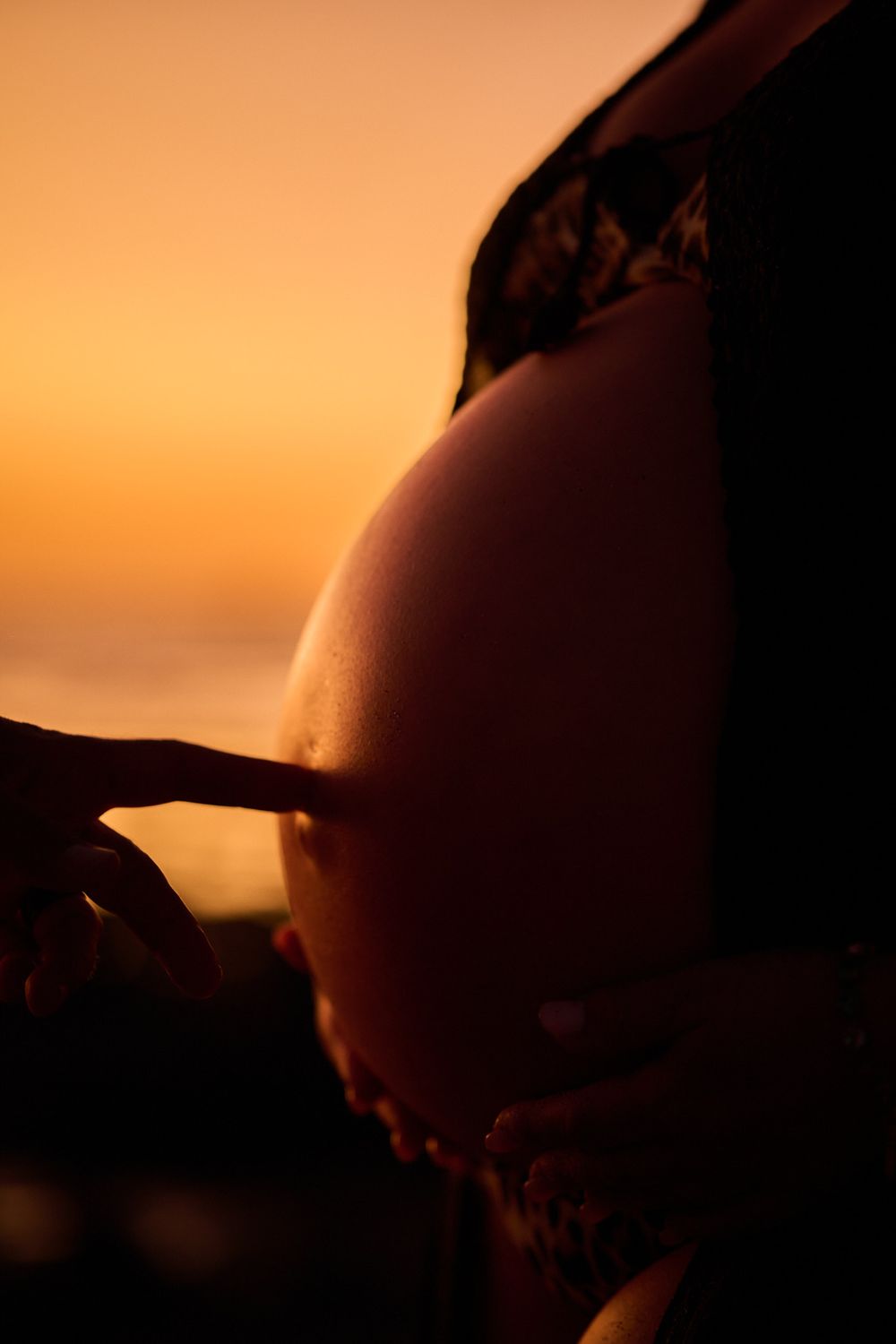 Silhouette of pregnant woman's belly with finger pointing at it against warm golden sunset sky.