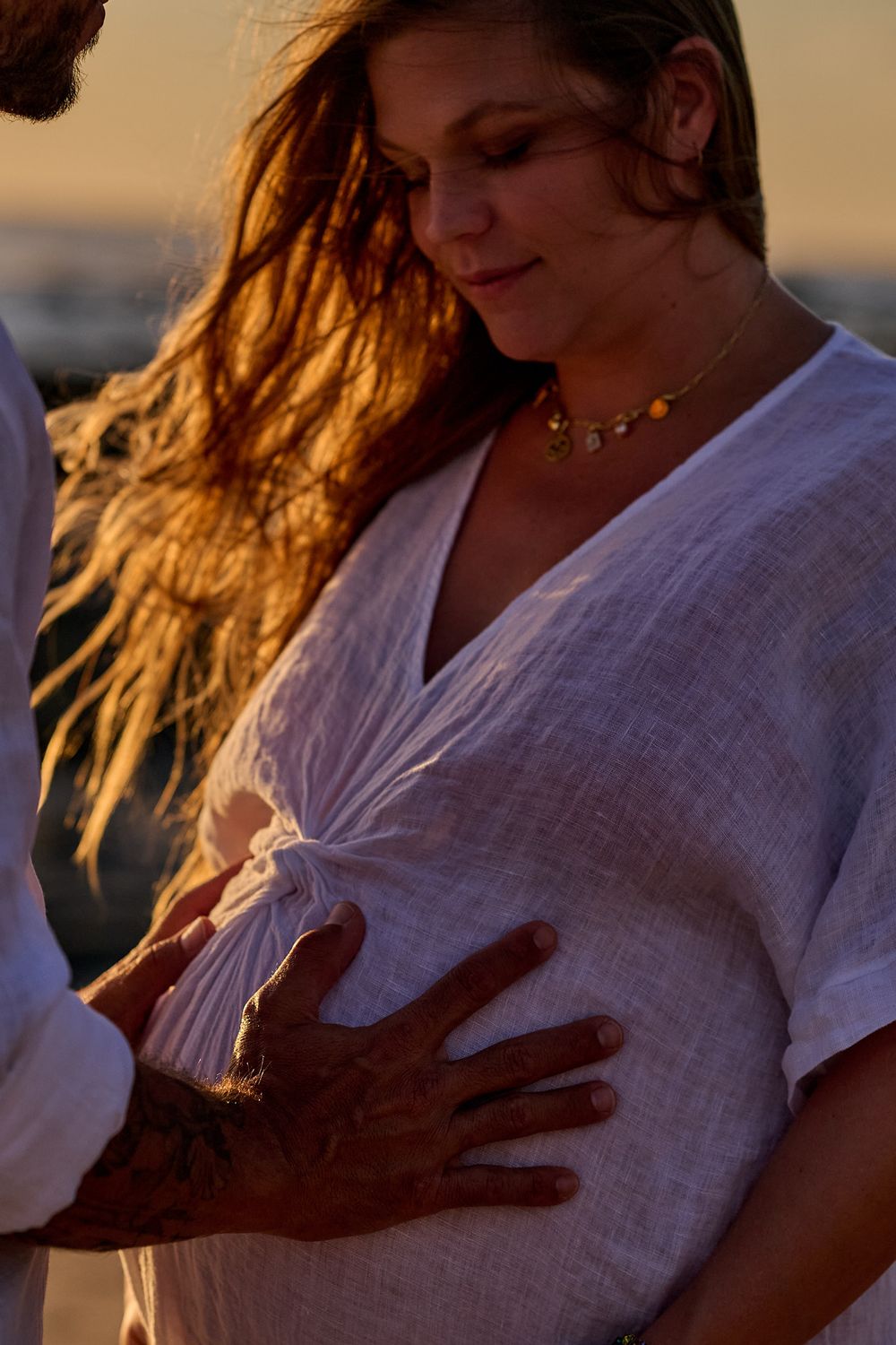 Pregnant woman in white linen dress touched by partner at golden sunset on beach.