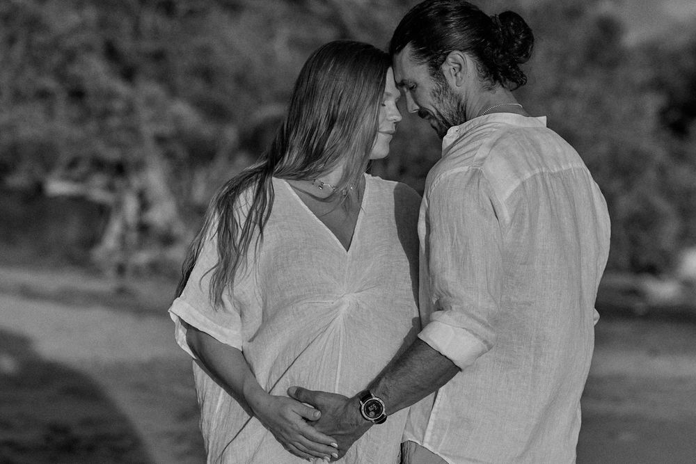 Black and white photo of a couple sharing a tender moment, woman in white dress with baby bump, man in linen shirt.