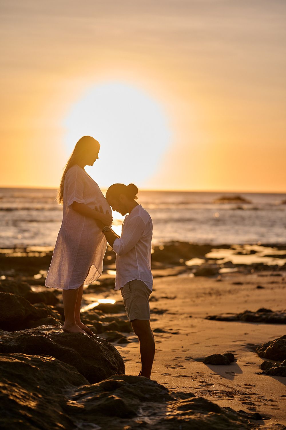Silhouette of pregnant couple standing on rocks at golden sunset beach, man leaning toward baby bump.