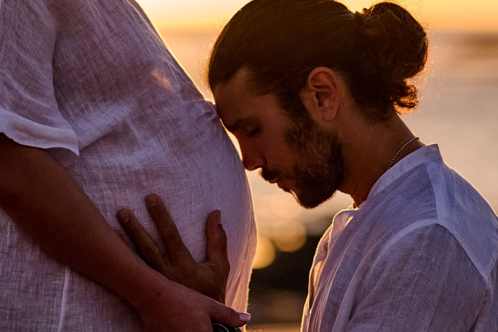 Man kneeling at sunset, gently touching pregnant woman's belly in a tender maternity moment outdoors.