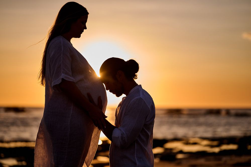 Silhouette of pregnant woman and partner sharing tender moment on beach at golden sunset by the ocean.