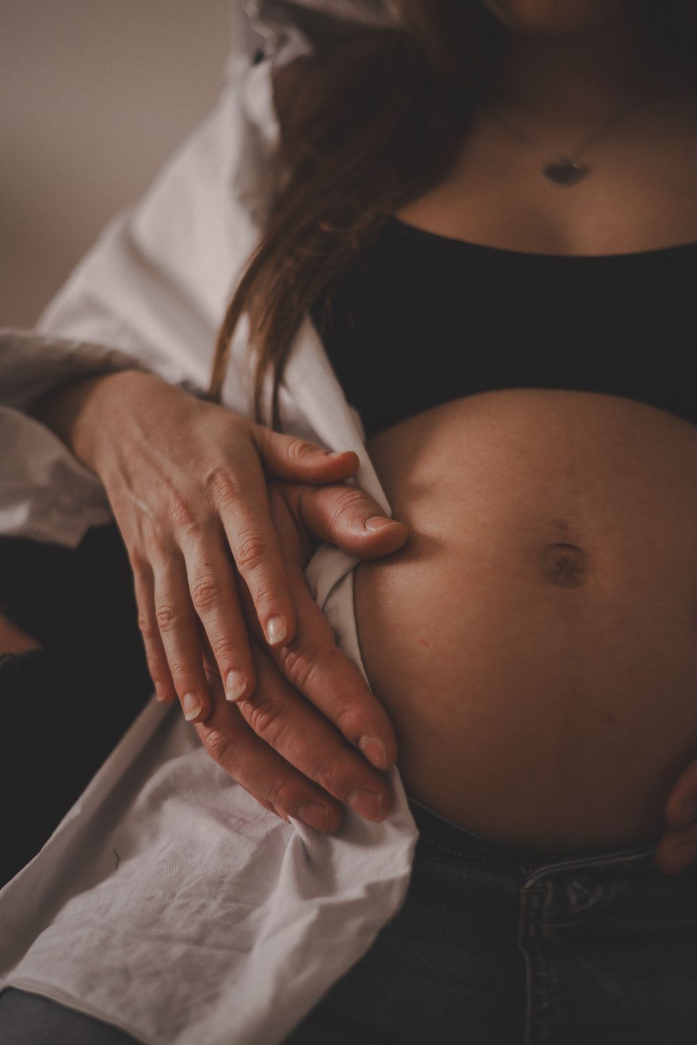Close-up of gentle hands resting on bare pregnant belly with white open shirt.
