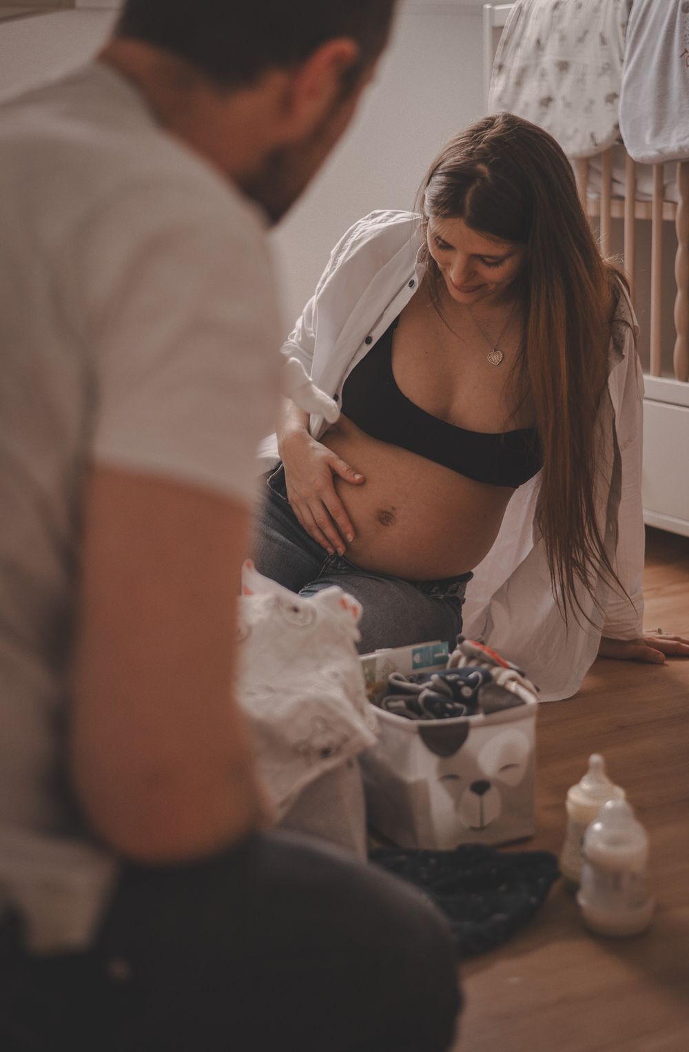 Pregnancy photo session showing couple sitting on wooden floor with baby supplies.
