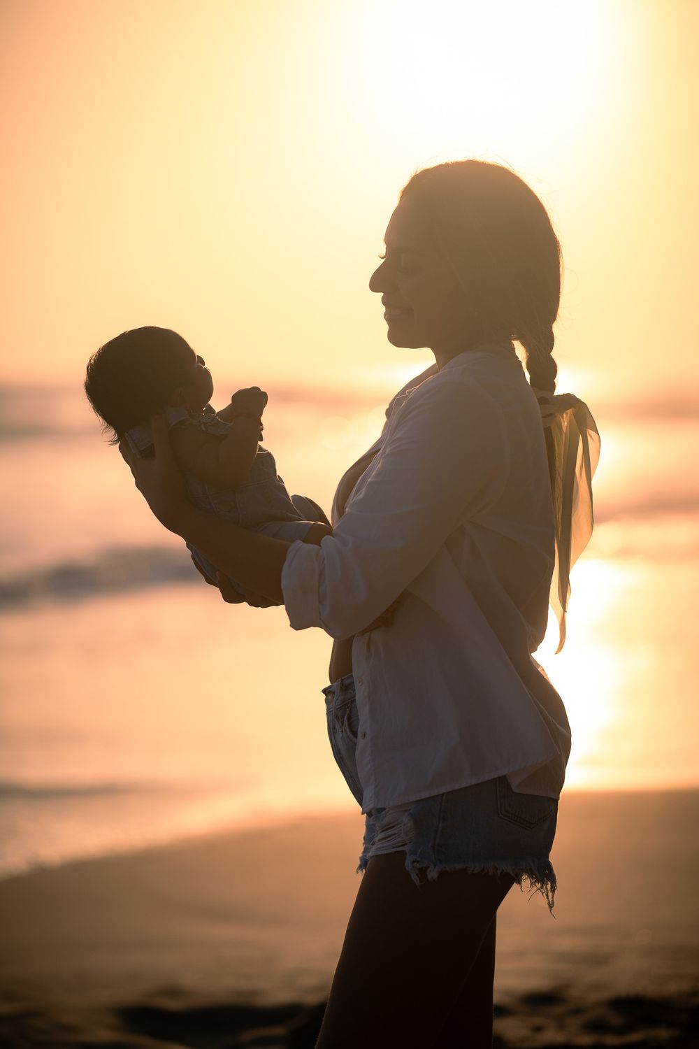 Silhouette of caregiver holding baby against golden sunset at beach.