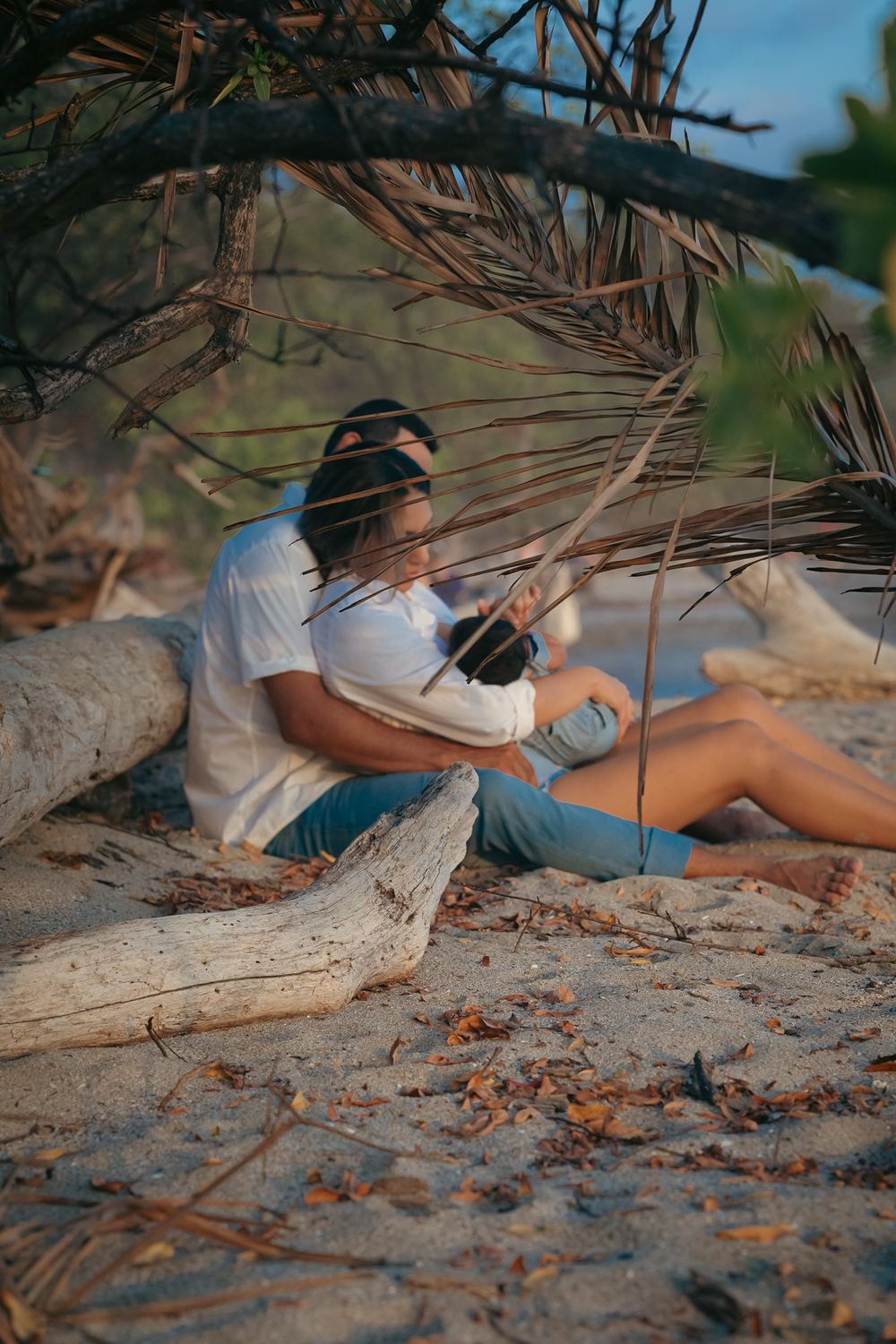 Person sitting with baby under palm fronds on sandy beach at sunset.