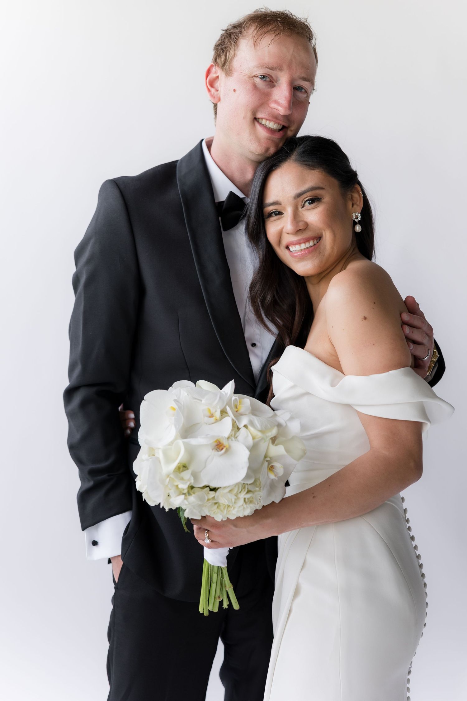 A bride in a white dress holds a bouquet of white orchids while posing with her groom in a black suit.