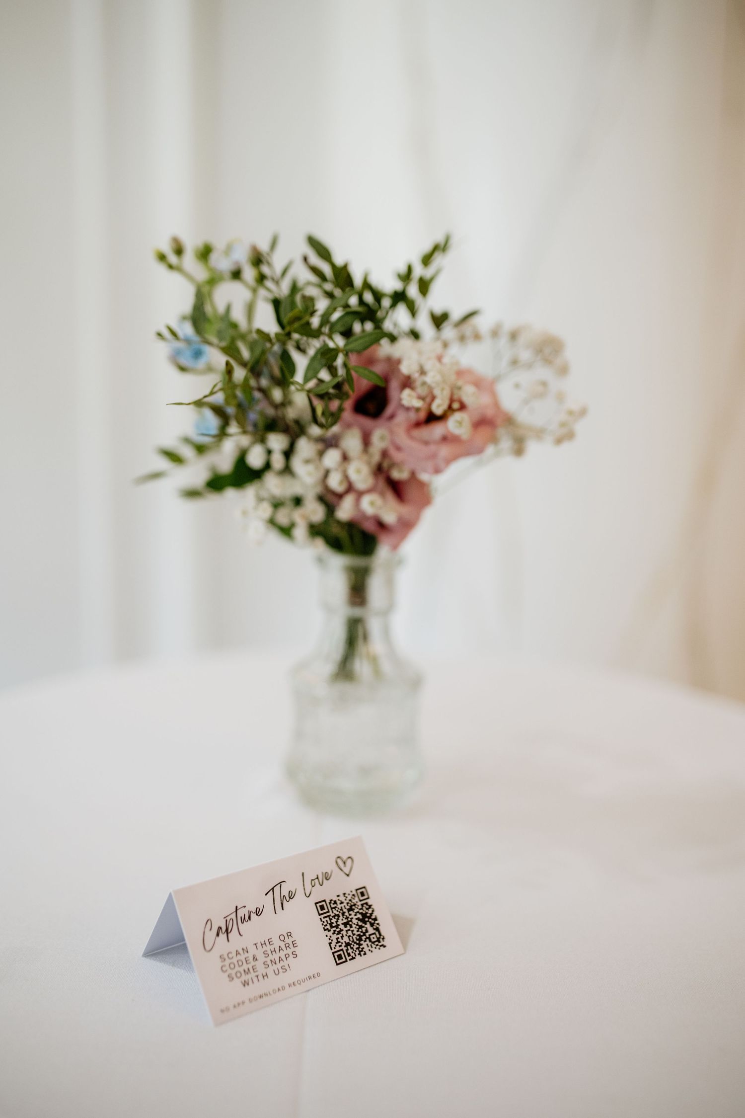 Elegant floral arrangement in a clear vase with pink roses and greenery next to a QR code place card.