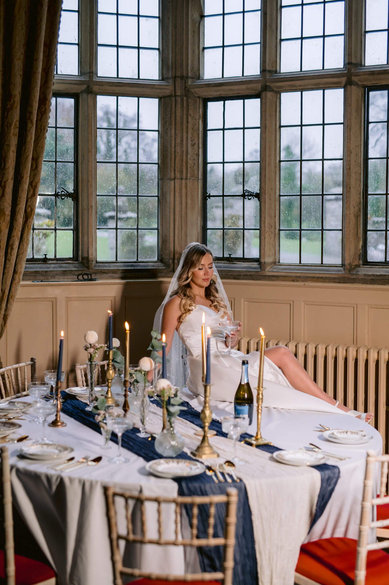 Elegant white and navy dining table setup with candlesticks and champagne in a sunlit room with large windows, bride posing.