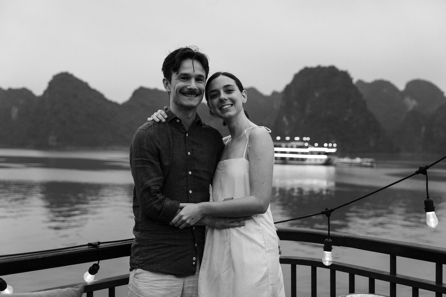 A black and white photo of a couple embracing on a boat with karst mountains and a lit cruise ship in distance.