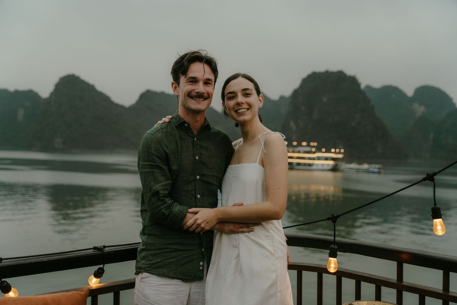 A couple shares a tender moment while standing on a boat deck with dramatic mountain scenery behind them.