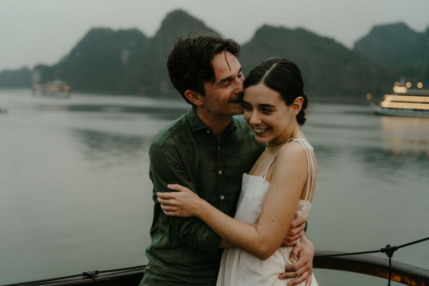 A couple shares a joyful moment on a boat deck with dramatic limestone cliffs and a cruise ship in view.