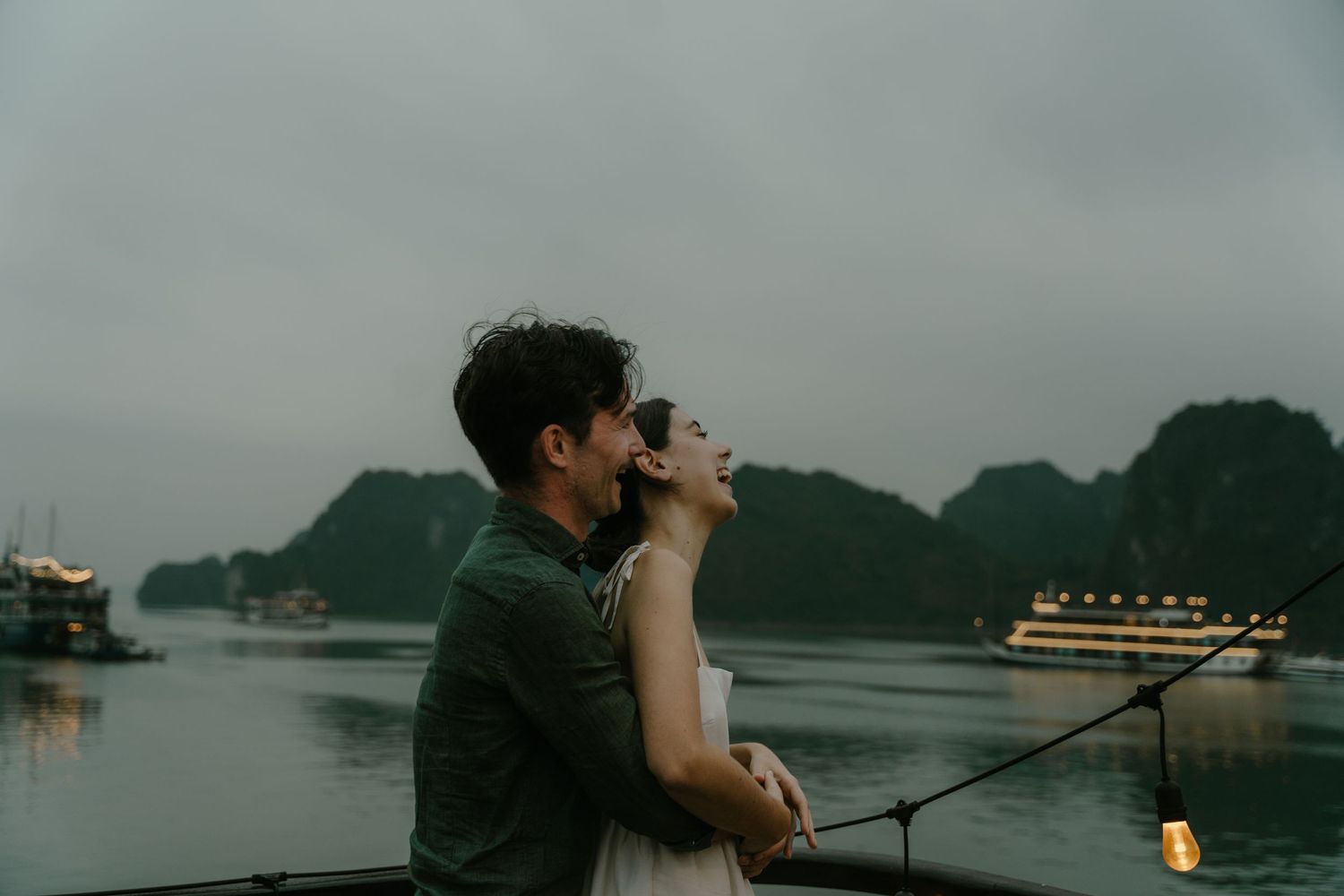 A couple embraces on a boat deck at dusk with dramatic karst mountains and illuminated cruise ships behind them.
