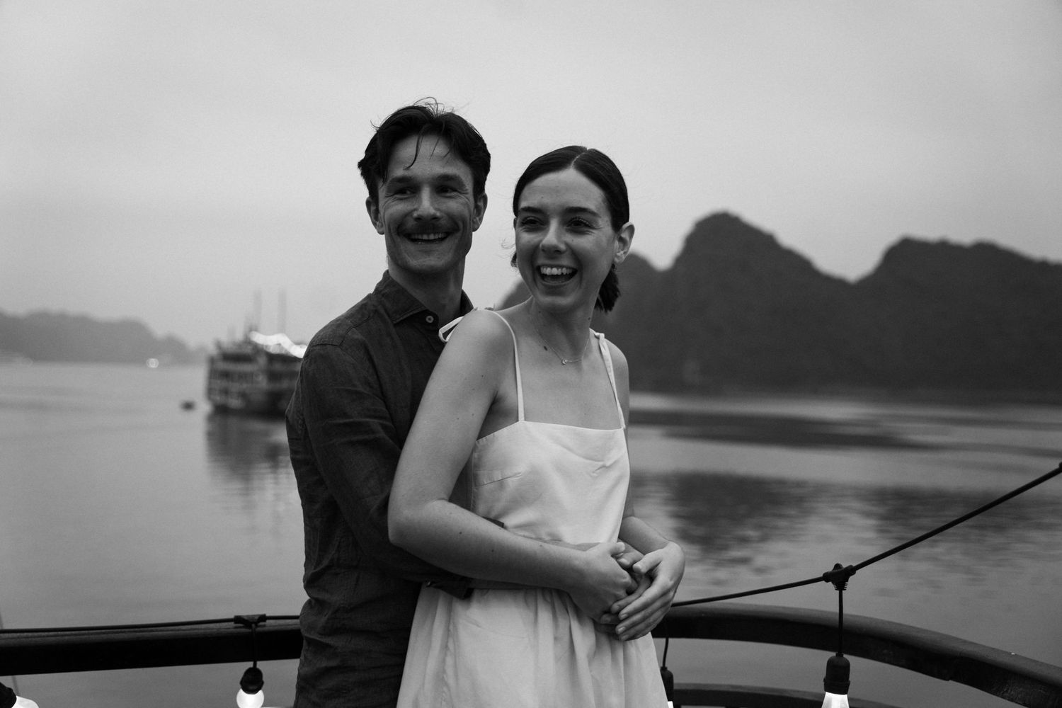 A black and white photo captures an intimate moment between a couple on a boat with mountains in background.