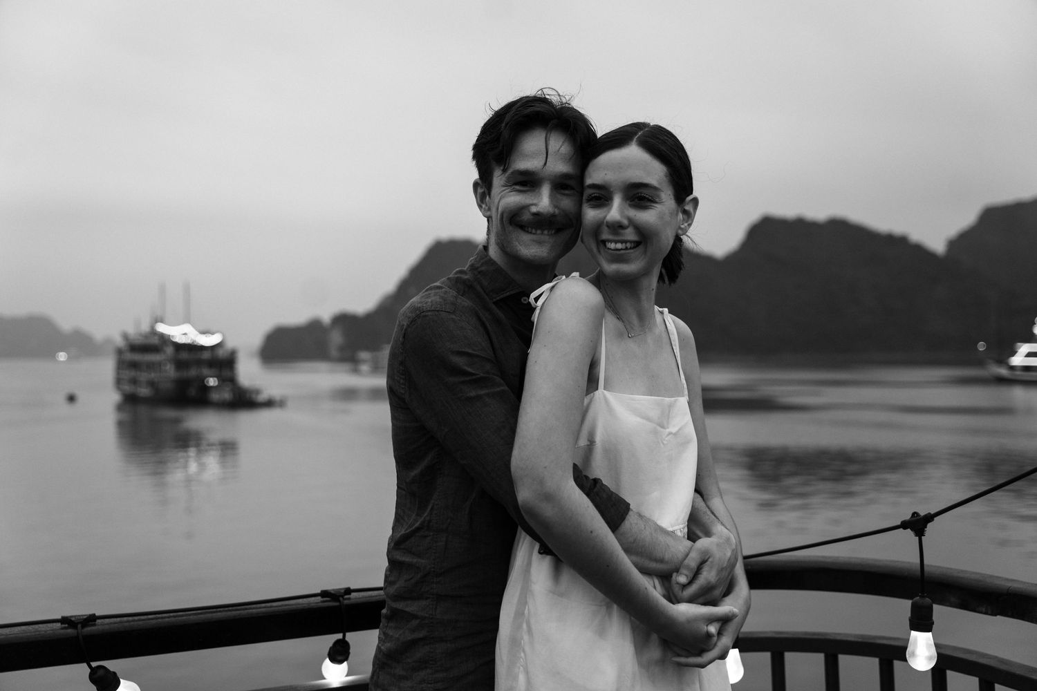 A black and white photo shows a couple in a loving embrace on a boat with mountains and water in background.