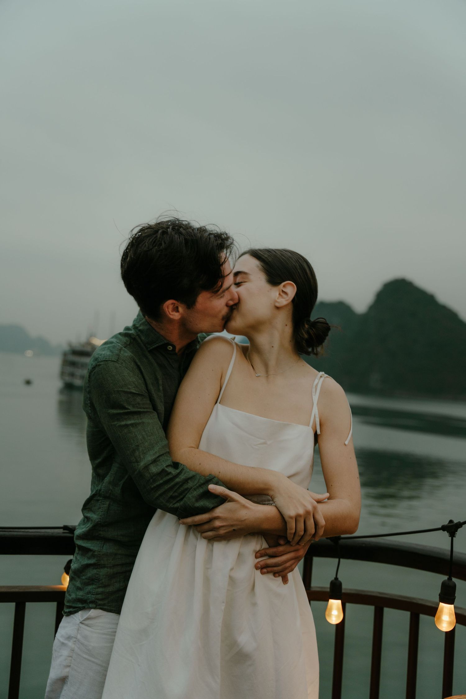 Romantic couple sharing an intimate kiss on a boat deck with misty mountains and string lights in the background.