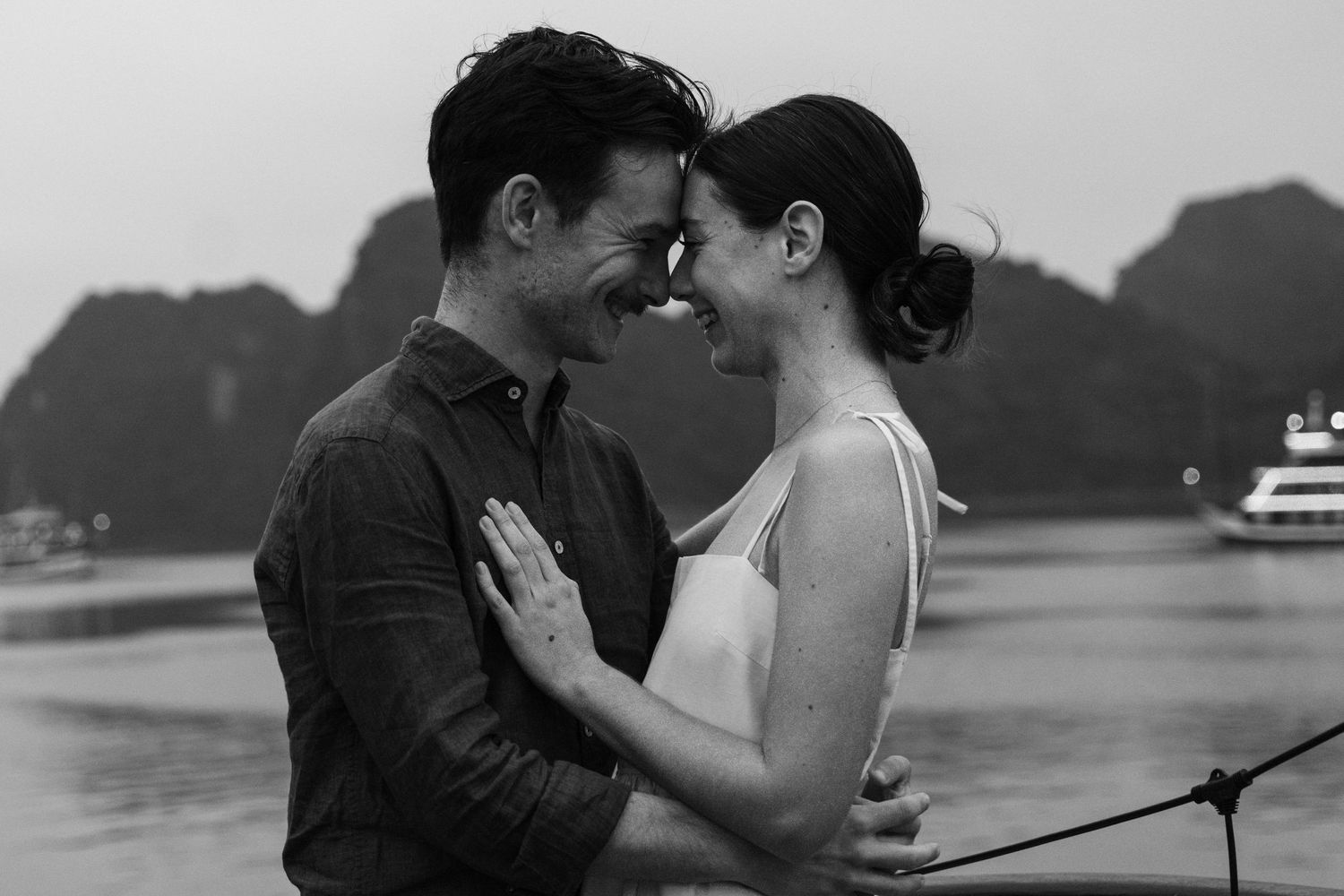 Black and white photo of a couple embracing against a backdrop of misty mountains and water.