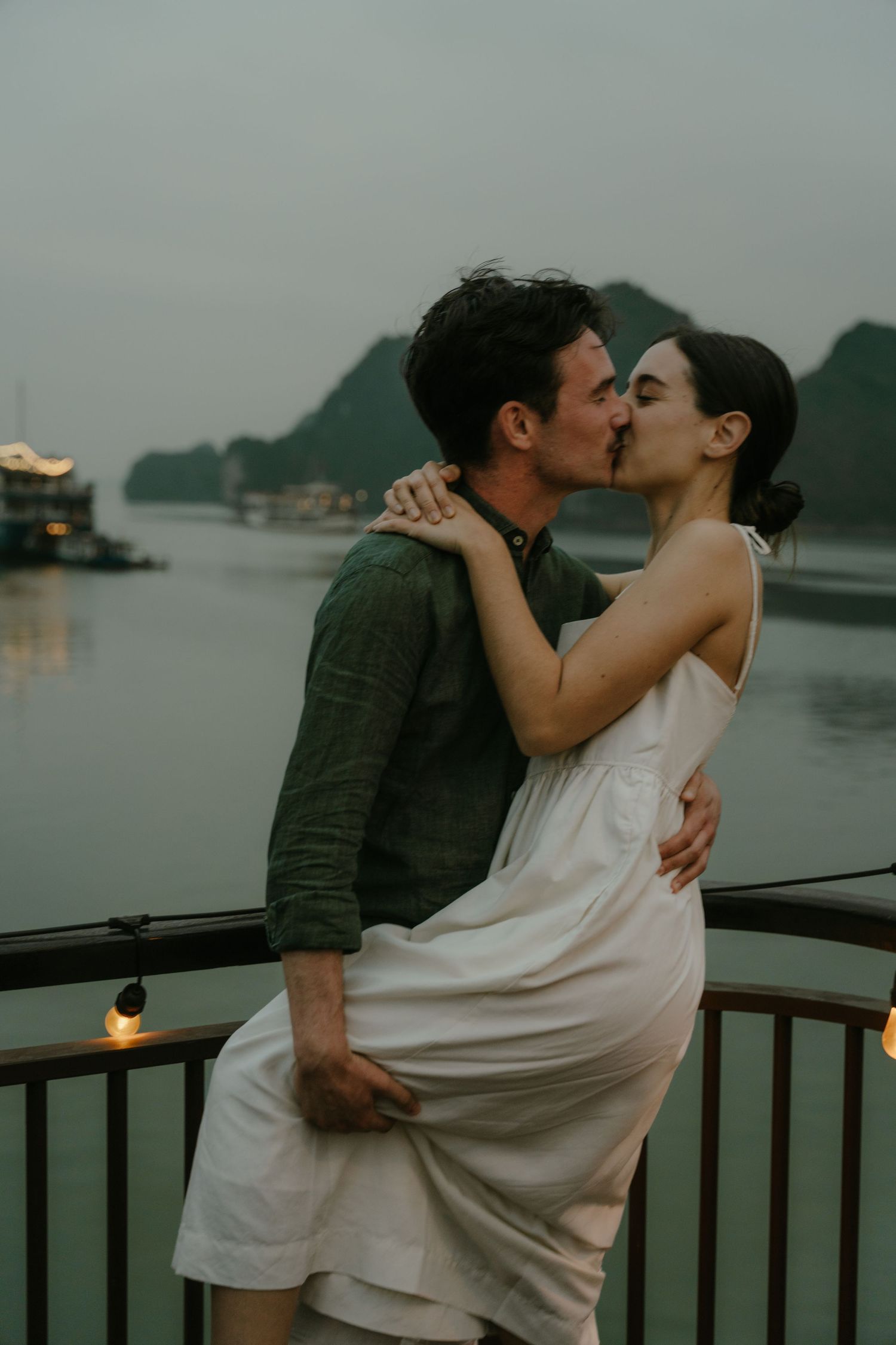 Couple in romantic embrace on boat deck at dusk with string lights and mountains in background.