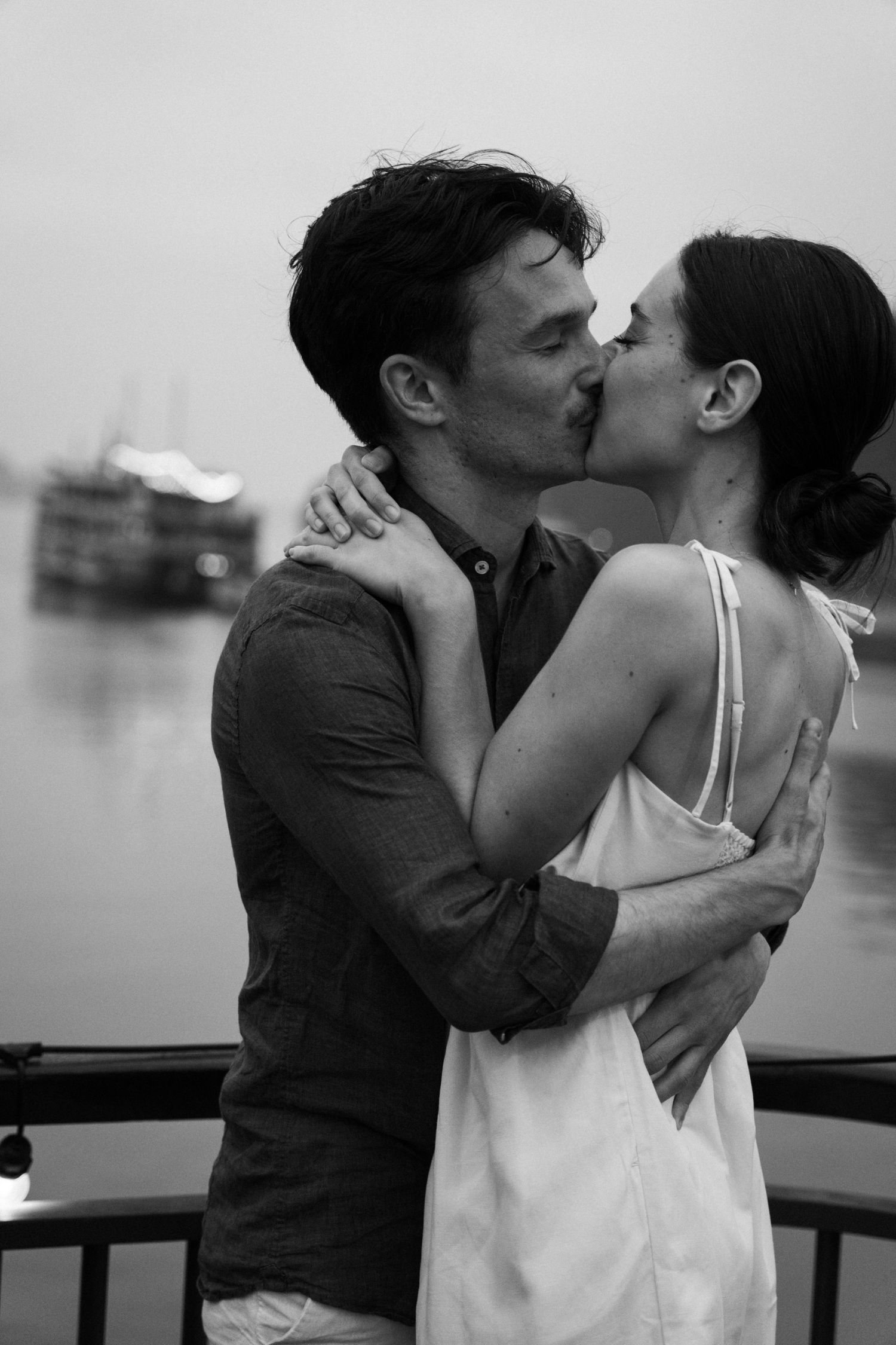 Black and white romantic moment captured of couple kissing on boat with water in background.